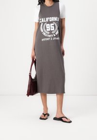 Robe grise, sans manches avec des manches t-shirt blanches, comportant le texte imprimé "California" et des détails graphiques. Assortie avec des tongs noires et un sac bordeaux.