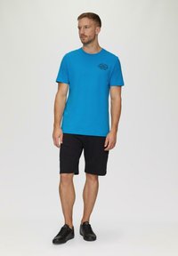 Turquoise katoenen T-shirt met korte mouwen, met een klein zwart logo op de linkerborst; gecombineerd met zwarte shorts en zwarte sneakers.