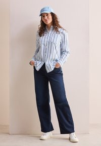Camisa de botones de rayas en azul claro con mangas largas y bolsillo frontal; combinada con jeans rectos en azul oscuro y zapatillas blancas.