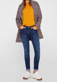 Multicolored houndstooth coat, bright mustard sweater, blue skinny jeans, white sneakers with brown accents, casually styled with hands in pockets.