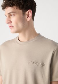 Šviesiai ruda medvilninė marškinėliai su apvalia kaklo iškirpte ir subtiliu Calvin Klein logotipu, išsiuvinėtu kairėje krūtinės pusėje. Lygus paviršius, laisvas siluetas.