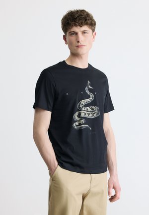 Unge mand med krøllet hår iført sort t-shirt med stort grafik af en snoet slange og beige bukser, stående med den ene hånd i lommen.