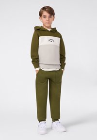 Niño de pie con las manos en los bolsillos, vistiendo una sudadera Champion verde oliva y beige, pantalones a juego y zapatillas blancas sobre un fondo plano.