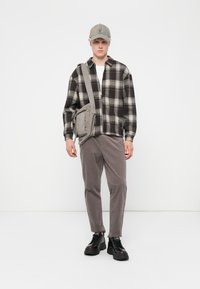 Schwarz-weiß kariertes Hemd, graue Cordhosen, graue Schultertasche und dunkle Schuhe. Accessoires umfassen eine graue Mütze. Schlichtes, modernes Design.