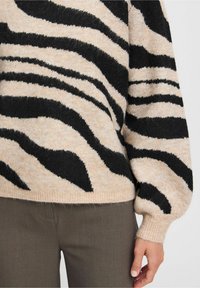 Pull en tricot beige avec de larges rayures zèbre noires, dotée d'une coupe décontractée et de manches bouffantes. Texture douce avec un ourlet côtelé.