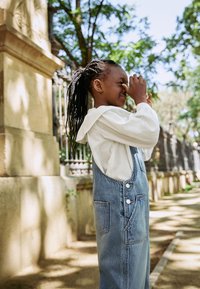 Salopette en denim avec une poche, de couleur bleu clair, portée par-dessus un t-shirt blanc texturé à manches longues avec une capuche. Enfant dans un environnement extérieur feuillu.