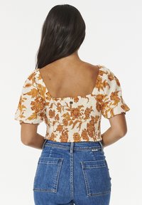Femme aux longs cheveux noirs portant un crop top à manches bouffantes floral orange et blanc et un jean taille haute bleu, vue de dos.