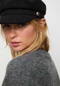 Cappello di lana nera con dettaglio in bottone dorato; il modello indossa un maglione grigio con texture. Un trucco morbido e naturale evidenzia le lentiggini sul viso.