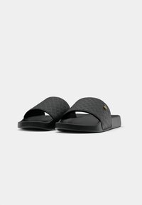 Une paire de sandales noires à glisser avec des brides larges texturées et un petit emblème logo doré sur un fond blanc.