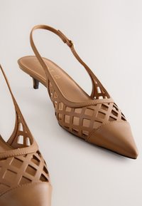 Escarpins slingback en cuir marron avec bouts pointus et motif ajouré en treillis sur les côtés, sur un fond uni.
