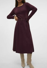 Burgundin midimekko, jossa on pitkät hihat, ribbed-tekstuuri, korkea vyötärö ja laskostettu helma; yhdistetty korkeisiin beigeisiin saappaisiin.