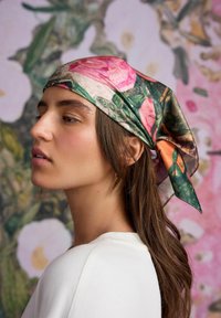 Hedvábný šátek s květinovým vzorem v růžových a zelených tónech, uvázaný vzadu. Modelka má na sobě bílý top, na pozadí je rozostřené květinové pozadí.