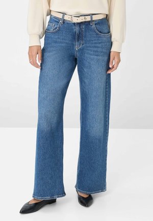 Femme portant un jean bleu droit avec une ceinture beige, un haut crème à manches longues et des ballerines noires à bout pointu, debout sur un sol blanc.