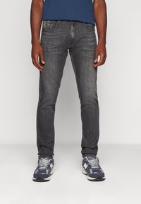 Uomo che indossa jeans slim-fit grigio scuro e sneakers blu, in piedi contro uno sfondo neutro.