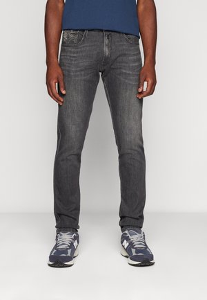 Uomo che indossa jeans slim-fit grigio scuro e sneakers blu, in piedi contro uno sfondo neutro.