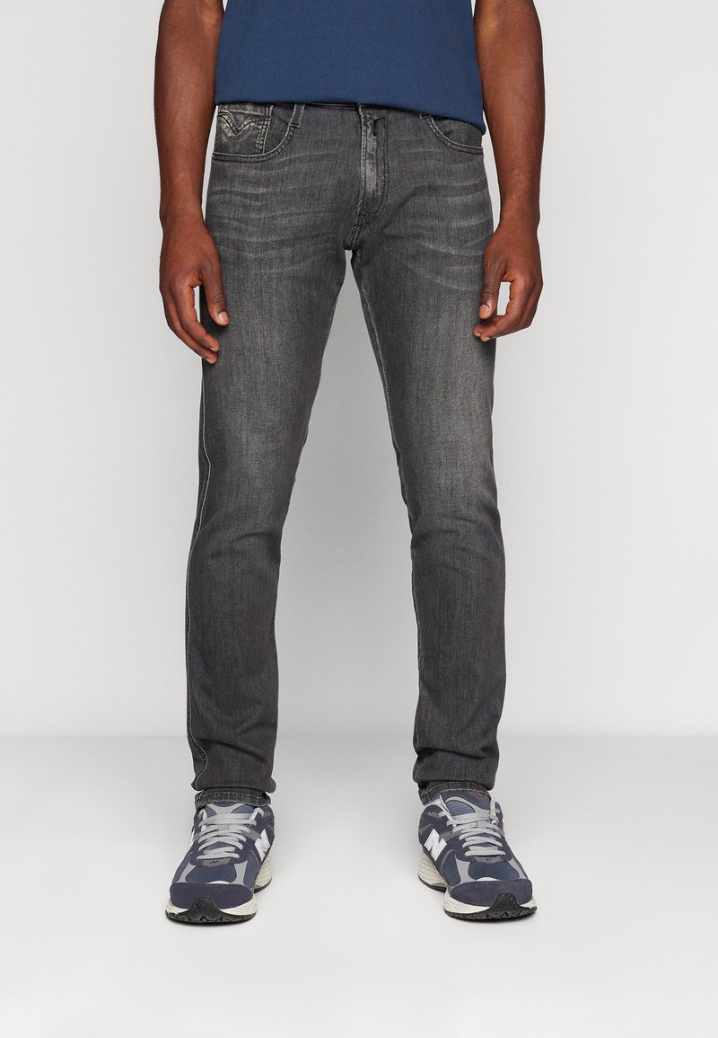 Uomo che indossa jeans slim-fit grigio scuro e sneakers blu, in piedi contro uno sfondo neutro.