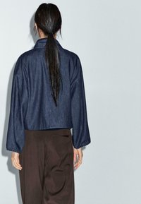 Persona con una lunga coda di cavallo scura che indossa una giacca blu scuro larga e pantaloni marroni a gamba larga, in piedi contro uno sfondo chiaro uniforme.