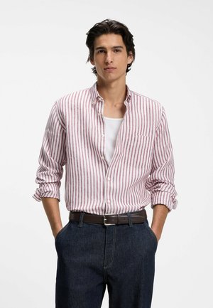 Młody mężczyzna o ciemnych włosach, noszący białą i czerwoną koszulę w paski na białym podkoszulku, z rękami w kieszeniach.