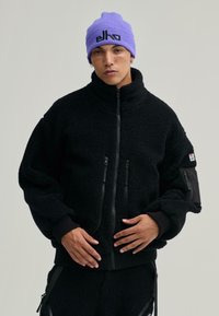 Czarna polarowa kurtka z wysokim kołnierzem, zapinana na zamek z przodu, z dwoma kieszeniami zapinanymi na zamek; ściągacze przy mankietach i u dołu; w połączeniu z jasnofioletowym czapką.