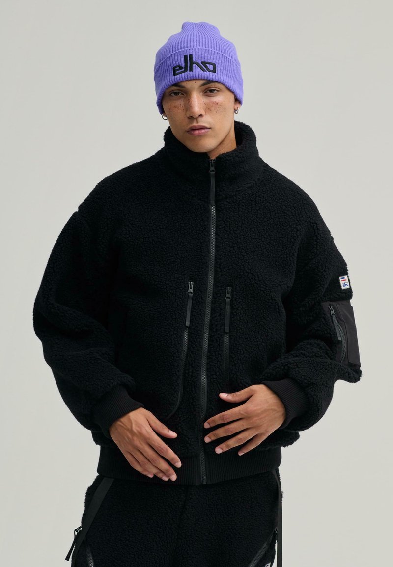 Czarna polarowa kurtka z wysokim kołnierzem, zapinana na zamek z przodu, z dwoma kieszeniami zapinanymi na zamek; ściągacze przy mankietach i u dołu; w połączeniu z jasnofioletowym czapką.