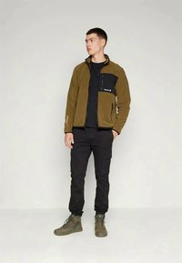 Giacca in pile con corpo marrone e tasca sul petto nera, chiusura con zip e dettaglio del logo. Abbinata a pantaloni cargo neri e stivali verdi.