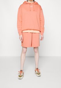 Persikofärgad sweatshirt med huva och framlogotyp, parat med matchande shorts. Lätta sneakers med orange snören och krämfärgade detaljer.