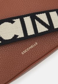 Ruskea teksturoitu nahkainen laukku, jossa on kultainen "COCCINELLE"-logo ja musta-valkoinen kudottu olkahihna, jossa on rohkeita geometrisia kuvioita.