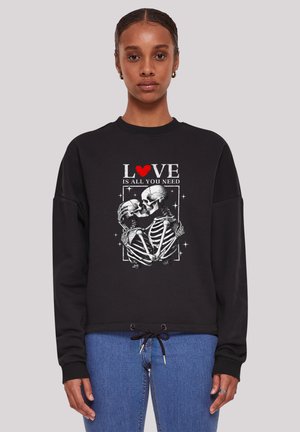 Frau trägt schwarzen Pullover mit Grafik von zwei sich küssenden Skeletten und dem Text „LOVE is all you need“, wobei das „O" durch ein rotes Herz ersetzt ist.