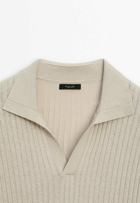 Beige ribbattu neulepolo, jossa on V-mallinen kaulus ja lyhyet hihat. Sisältää hienovaraisen tekstuurin ja tikkaukset kauluksen kohdalla.