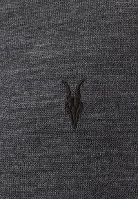 Dunkelgrauer gestrickter Stoff mit einer subtilen Textur, ausgestattet mit einem kleinen schwarzen gestickten Ziegenschädel-Logo, das sich oben links befindet.