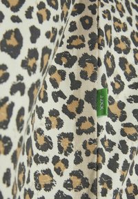 Textilie s leopardím potiskem s černými a hnědými skvrnami na krémovém pozadí. Na okraji je viditelná zelená etiketa, materiál je texturovaná plátno.