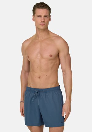 Uomo muscoloso con capelli pettinati all'indietro che indossa pantaloncini da bagno blu con coulisse, in piedi davanti a uno sfondo chiaro e uniforme.