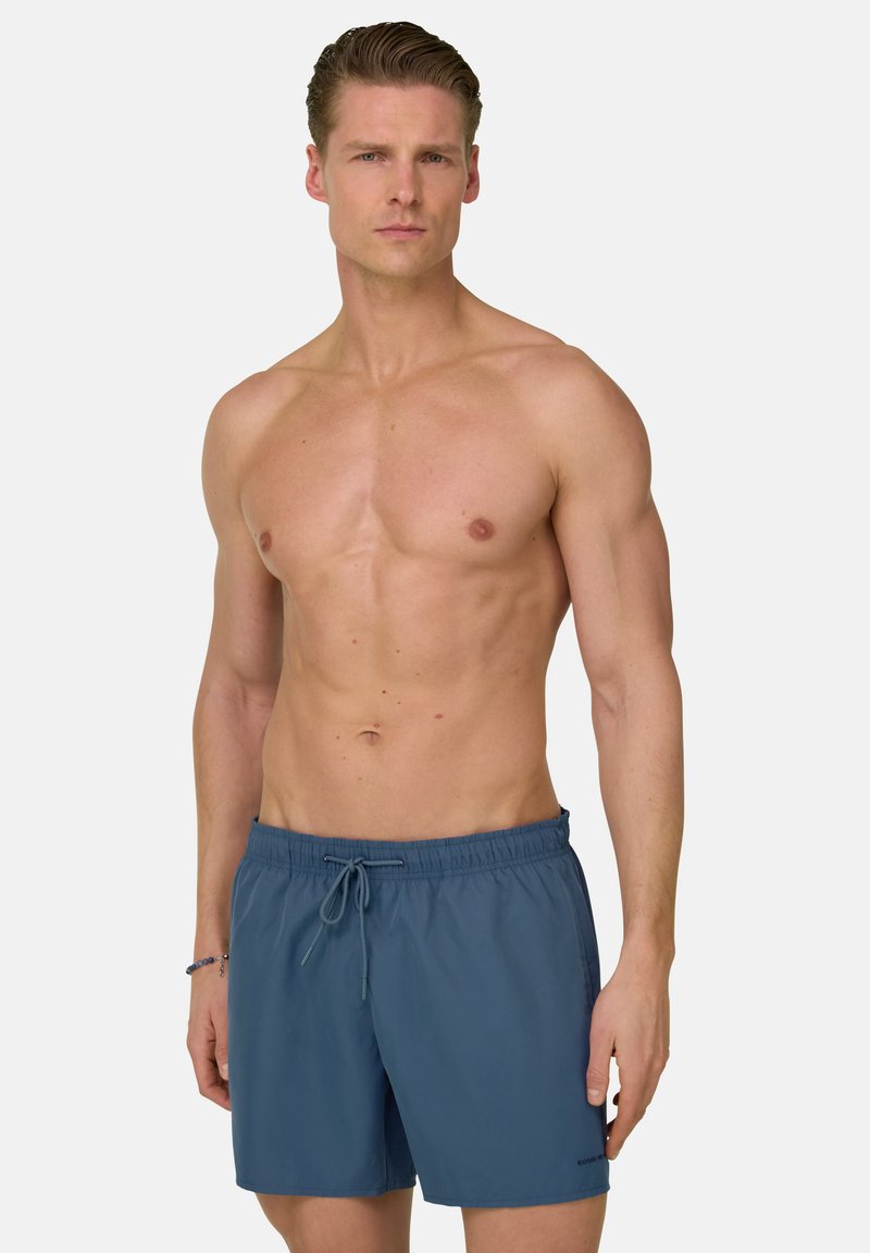 Homme musclé aux cheveux plaqués en arrière, portant un short de bain bleu à cordon, debout devant un fond clair uni.