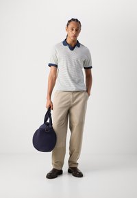 Polo à manches courtes rayé en blanc et marine, associé à un pantalon cargo beige. Tient un sac rond marine, porte des bottes sombres.