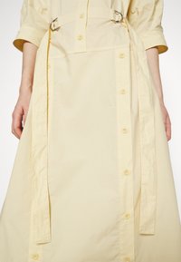 Lång gul kn äppt klänning med korta ärmar, föreskriven med en sidöppning och två runda metallringar för justering. Slätt, lätt tyg.