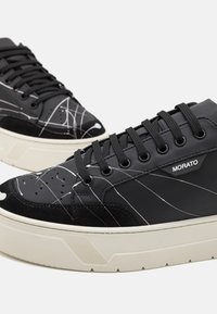 Fekete sneaker bőr és velúr keverékből, fehér vonalmintákkal, kerek orral, vastag krémszínű talppal és fekete fűzőkkel.