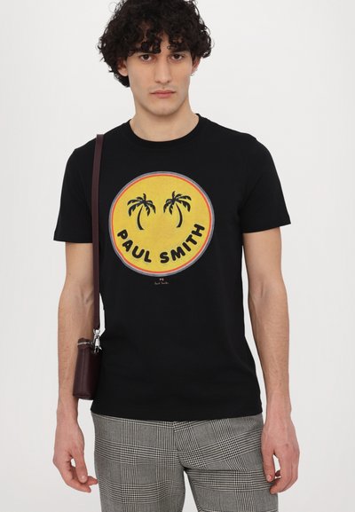 T-shirt en coton noir avec un graphique rond jaune représentant des palmiers et le texte "PAUL SMITH", porté avec un pantalon à carreaux gris et un sac marron.