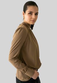 Vrouw met donker haar die gouden hoepeloorbellen en een bruine lange mouw hoge hals top draagt, staand tegen een effen lichtgrijze achtergrond.