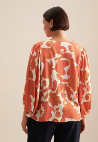 Blusa floreale color crema con grande motivo corallo e verde, dotata di maniche arricciate e una vestibilità ampia, abbinata a pantaloni scuri.