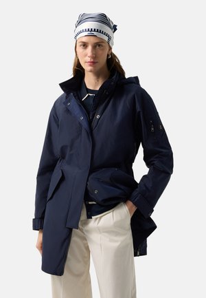 Frau trägt eine dunkelblaue Jacke, gestreiften Kopfschleier, dunkles Oberteil und cremefarbene Hose, steht mit einer Hand in der Tasche vor einem schlichten Hintergrund.