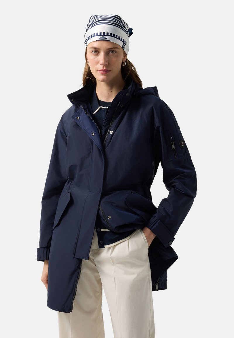 Vrouw die een marineblauwe jas, gestreepte hoofddoek, donker shirt en crème broek draagt, staand met één hand in de zak tegen een effen achtergrond.