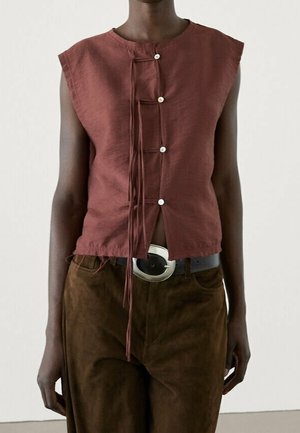 Ærmeløs top i rustfarve med asymmetrisk knaplukning og bindedetalje, kombineret med mørkebrune bukser og et sort bælte med sølvspænde.