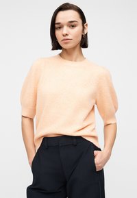 Femme aux cheveux courts et foncés, portant un pull à manches courtes pêche et un pantalon noir, les mains dans les poches, debout devant un fond clair uni.