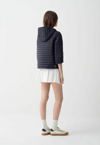 Marine pufferjack met een capuchon en korte mouwen, met een gequilt textuur. Draag ik over een witte geplooide rok en sneakers met donkere accenten.