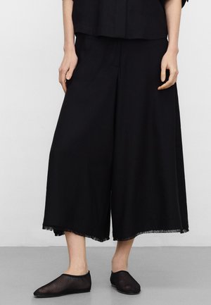 Person trägt schwarze, weit geschnittene, kurz geschnittene Hose mit rohen Säumen und schwarze Mesh-Slipper, steht vor einem schlichten Hintergrund.