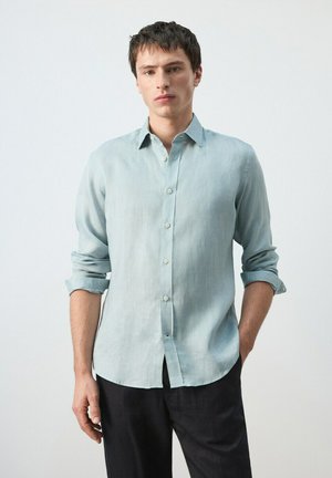 Joven con camisa azul clara de botones y mangas remangadas, pantalones oscuros, de pie frente a un fondo blanco liso.