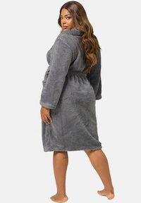 Kvinna med långt hår som bär en grå plyschrock knuten i midjan, står barfota och tittar över axeln mot en vit bakgrund.