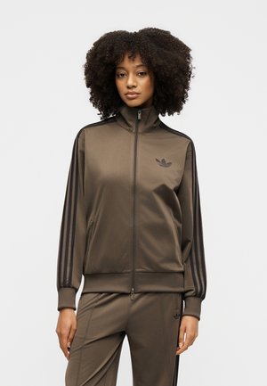 Frau mit lockigem Haar, die einen braunen Adidas-Trainingsanzug mit schwarzen Streifen trägt und vor einem schlichten hellen Hintergrund steht.