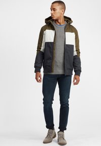 Green, white, and navy zip-up jacket with hood; knit gray sweater underneath; dark jeans; light gray ankle boots. Neutral background.