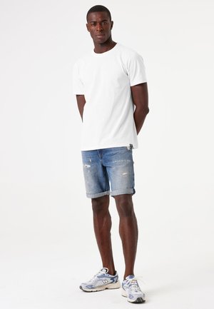 Witte t-shirt met korte mouwen, gecombineerd met versleten blauwe denimshorts met opgerolde zomen en grijze sneakers met witte accenten.